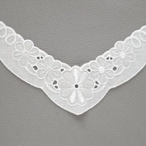 Könnte beinhalten: Ein weißer, bestickter Spitzenkragen mit floralem Design. Der Kragen hat einen gewellten Rand und eine V-Ausschnitt-Form. Die filigrane Stickerei umfasst Blumen und Durchbrucharbeiten, ideal, um Kleidung einen Vintage-Touch zu verleihen.