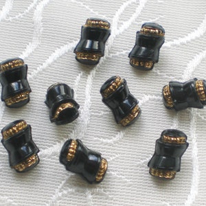 Può includere: Dieci bottoni neri e dorati con un design decorativo. I bottoni hanno la forma di un piccolo barile con un bordo dorato attorno al centro.