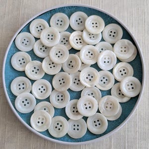 Peut inclure: Ensemble de boutons blancs ronds, chacun percé de quatre trous, disposés dans un récipient bleu circulaire. Les boutons présentent un aspect nacré, suggérant de la nacre. Le récipient a un bord blanc.