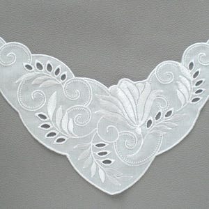 Könnte beinhalten: Weiße Spitzenkrause mit zartem Blumendesign. Die Krause ist aus einem durchsichtigen Stoff gefertigt und hat einen gewellten Rand.