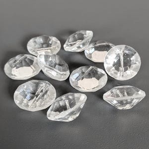 Könnte beinhalten: Eine Sammlung klarer, facettierter Acrylperlen. Die Perlen sind oval und rund mit flacher Rückseite und einem Loch zum Auffädeln. Die Perlen sind auf einer grauen Oberfläche verstreut. Diese Perlen eignen sich für die Schmuckherstellung und Bastelarbeiten.