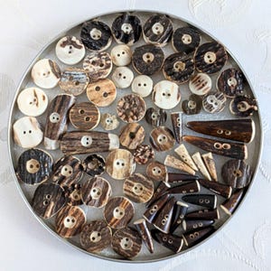 Pode incluir: Uma coleção de botões variados numa bandeja prateada. Os botões são feitos de materiais naturais, apresentando tons de castanho, creme e preto. Variam em tamanho e forma, incluindo designs redondos, ovais e em forma de chifre. Alguns têm dois ou quatro furos.