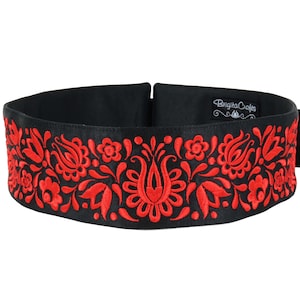 Op de afbeelding: Zwarte satijnen riem met een rood geborduurd bloemenmotief. De riem heeft een gespsluiting en is gelabeld "Brigita Crafts".