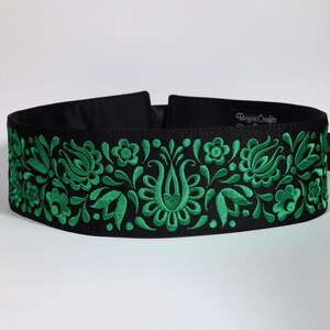 Puede incluir: Cinturón negro con bordado floral verde. El cinturón presenta un patrón detallado de flores y hojas estilizadas. El nombre de la marca "Brigitta Crafts" es visible en el cinturón.