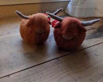 Può includere: Due ornamenti in feltro a forma di mucca delle Highlands. Ogni mucca ha una forma rotonda con lana marrone-arancio, corna grigie e un piccolo viso chiaro. Sono su una superficie di legno vicino a una finestra.