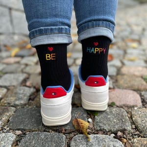 Könnte beinhalten: Ein Paar schwarze Socken mit bunten gestickten Herzen und den Worten "BE" und "HAPPY". Die Socken werden mit weißen Sneakers mit blauen und roten Akzenten getragen.