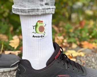 May include: White athletic socks with an embroidered avocado character wearing a headband and exercising. The text "Avecardio" is below the avocado. The socks are worn with black athletic shoes and gray jeans.