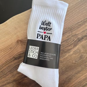 Op de afbeelding: Witte sokken met zwarte geborduurde tekst die "Welt bester PAPA" luidt, met een rood hart tussen de woorden "bester" en "PAPA". De sokken zijn verpakt met een zwart label waarop "LEDER STICH & CO" en "WO JEDER STICH EIN STATEMENT IST - INDIVIDUELL UND EINZIGARTIG." staat.