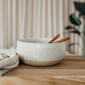 Könnte beinhalten: Eine gesprenkelte, weiße Keramikschüssel mit beigem Boden, in der sich zwei Holzkochlöffel befinden. Ein beiges Leinentuch liegt auf einer Holzoberfläche. Ein Küchenartikel zum Servieren von Salaten.