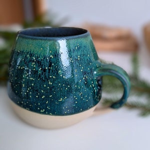 Può includere: Una tazza in ceramica verde e bianca con una glassa macchiettata. La tazza ha un corpo arrotondato e un manico.