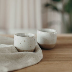 May include: Two white ceramic mugs with brown speckles sit on a light brown wooden surface covered with a beige linen cloth.