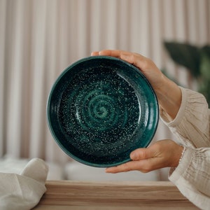 Könnte beinhalten: Eine tiefgrüne Keramikschüssel mit gesprenkeltem Glase und einem wirbelnden Muster. Die Schüssel wird von den Händen einer Person gehalten.