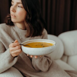 Peut inclure: Une personne tenant un bol blanc de soupe orange avec une cuill&egrave;re. Le bol a un gla&ccedil;age mouchet&eacute;.