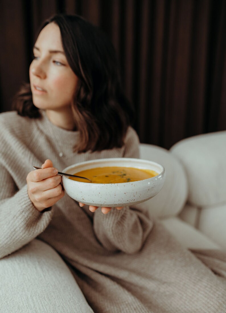 Peut inclure: Une personne tenant un bol blanc de soupe orange avec une cuill&egrave;re. Le bol a un gla&ccedil;age mouchet&eacute;.