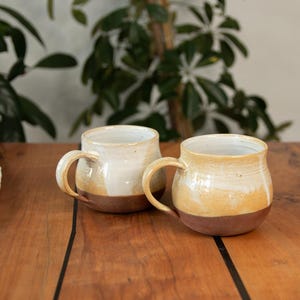 Könnte beinhalten: Zwei Keramikbecher mit weißem Überzug und braunem Rand. Die Becher stehen auf einem Holztisch.