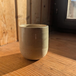 Könnte beinhalten: Beige Keramiktasse mit gesprenkeltem Design. Die Tasse hat eine zylindrische Form mit leicht abgerundetem Boden. Sie steht auf einer Holzoberfläche, mit Schatten.