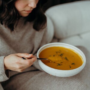 Peut inclure: Une personne est assise sur un canap&eacute; et mange un bol de soupe orange avec une cuill&egrave;re. La soupe est garnie d'herbes vertes.