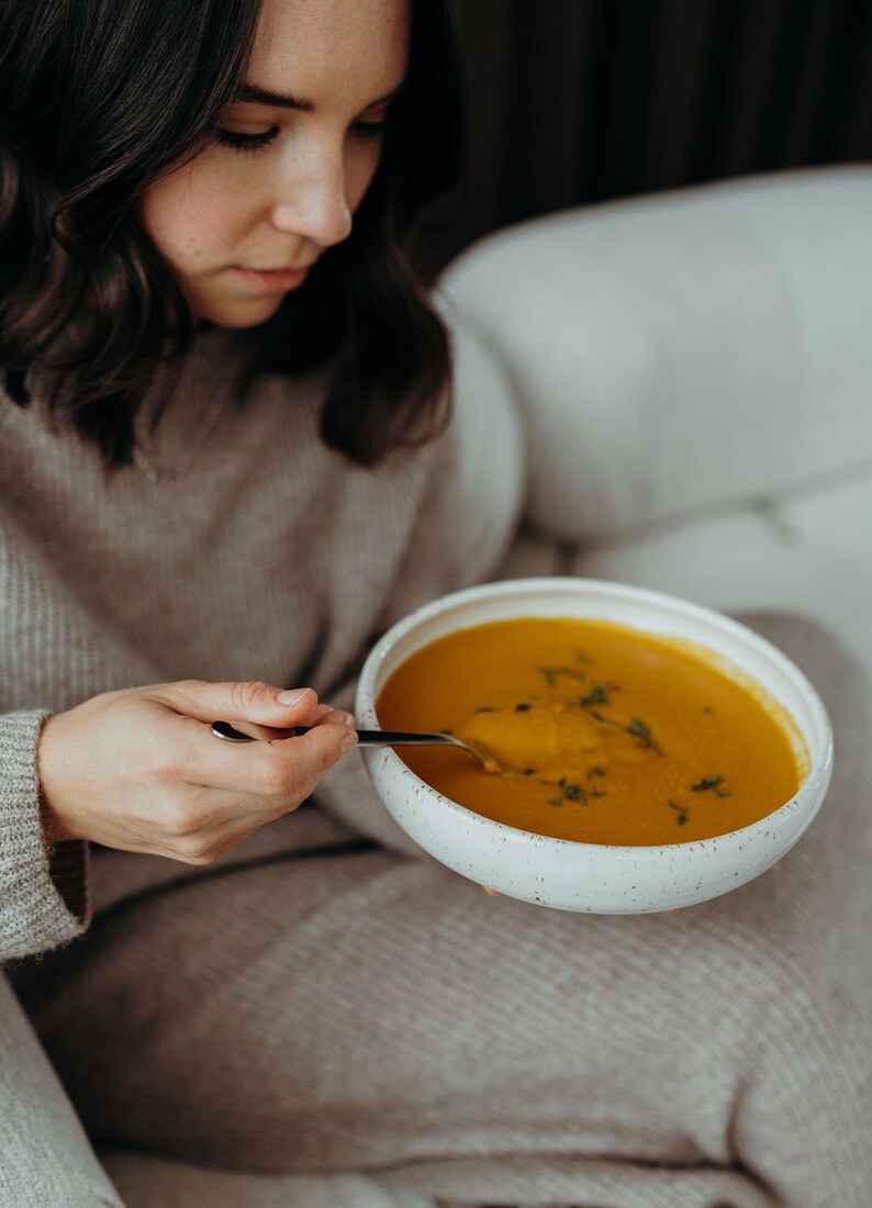 Peut inclure: Une personne est assise sur un canap&eacute; et mange un bol de soupe orange avec une cuill&egrave;re. La soupe est garnie d'herbes vertes.