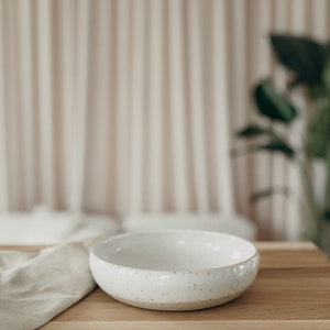 Peut inclure: Un bol en c&eacute;ramique blanche avec un gla&ccedil;age mouchet&eacute; pos&eacute; sur une table en bois. Le bol est vide et pr&ecirc;t &agrave; &ecirc;tre rempli.