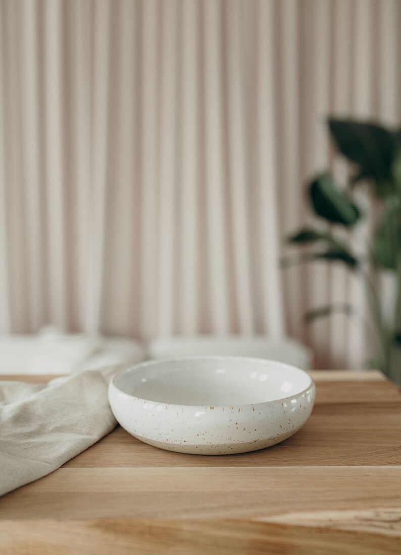 Peut inclure: Un bol en c&eacute;ramique blanche avec un gla&ccedil;age mouchet&eacute; pos&eacute; sur une table en bois. Le bol est vide et pr&ecirc;t &agrave; &ecirc;tre rempli.