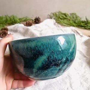 Op de afbeelding: Een diep groene keramische kom met een gespikkeld glazuur. De kom heeft een afgeronde vorm en een gladde, glanzende afwerking.