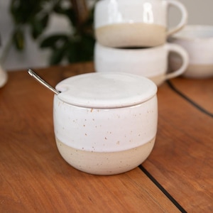 Könnte beinhalten: Eine weiße Zuckerdose aus Keramik mit Deckel und einem kleinen Löffel. Die Dose hat eine gesprenkelte Glasur und einen cremefarbenen Streifen in der Mitte.