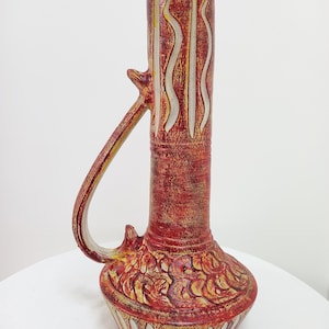 May include: A red and white ceramic vase with a unique design. The vase has a tall, cylindrical neck and a wide, rounded base. The neck is decorated with white wavy lines, and the base is decorated with a floral pattern.