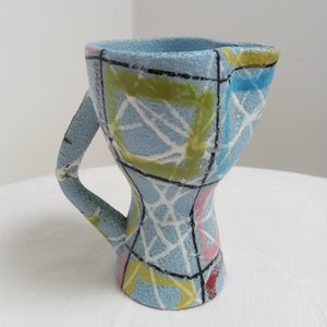May include: A blue ceramic pitcher with a geometric design in yellow, green, pink, and black. The pitcher has a handle and a wide base.
