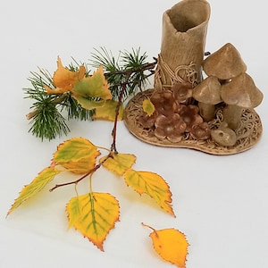 Könnte beinhalten: Eine Keramikvase mit natürlicher, erdiger Ästhetik, mit zylindrischer Form und einem Sockel, der mit geformten Pilzen und anderen Waldelementen verziert ist. Die Anordnung umfasst Zweige mit gelben und grünen Blättern.