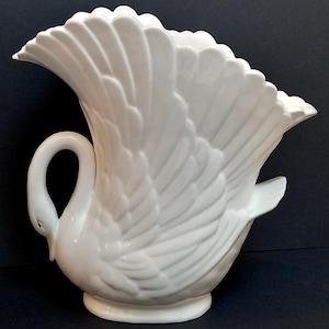 May include: A white ceramic swan vase. The vase has a curved neck and a flared, fan-shaped body. The design includes detailed feather textures and a small, round base. The swan's head forms a handle.
