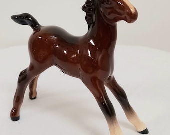 May include: A ceramic figurine of a brown foal with a glossy finish. The foal has a light brown face and lower legs, with black hooves and a black tail. The figurine is posed in a playful stance.