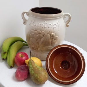 Könnte beinhalten: Eine weiße Keramikdose mit braunem Deckel und braunem Rand. Die Dose hat ein erhabenes Design aus Früchten und das Wort "Pantopf" auf der Vorderseite. Die Dose steht auf einer weißen Oberfläche mit Bananen, Äpfeln, einer Birne und einer Zitrone.