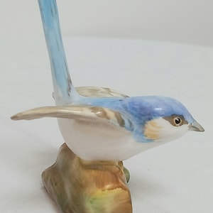 May include: A porcelain figurine of a bluebird perched on a brown and green rock. The bird has a long, thin tail and a white belly.