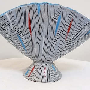Puede incluir: Un jarrón de cerámica gris en forma de abanico con un patrón geométrico negro, blanco, rojo y azul. El jarrón tiene una base ancha y plana.