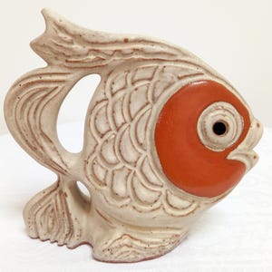Op de afbeelding: Een keramieken visfiguur met een gespikkeld beige lichaam, een gestructureerd schubpatroon en een glad, oranje cirkelvormig gebied rond het oog. Het beeldje heeft een opengewerkte staart en een kleine, open mond, afgewerkt met een matte finish.