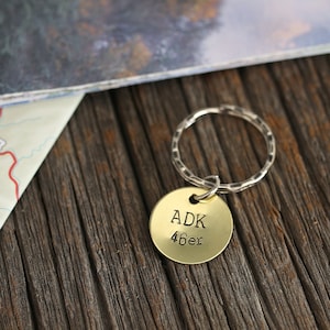 May include: A gold-colored keychain with a round tag that says "ADK 46er" on a wooden surface. The keychain is next to a folded map.