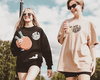 以下が含まれることがあります： 「Wild Leisure」とプリントされた黒とベージュのTシャツを着た2人。左の人は卓球ラケットを2本持っている。右の人は炭酸飲料の缶を持っている。