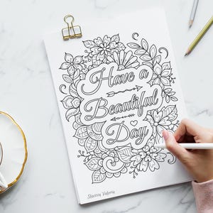 May include: A black and white floral coloring page with the words "Have a Beautiful Day" is held by a gold binder clip. A hand is coloring the page with a white pen. A cup of tea sits on a white saucer with a gold rim.