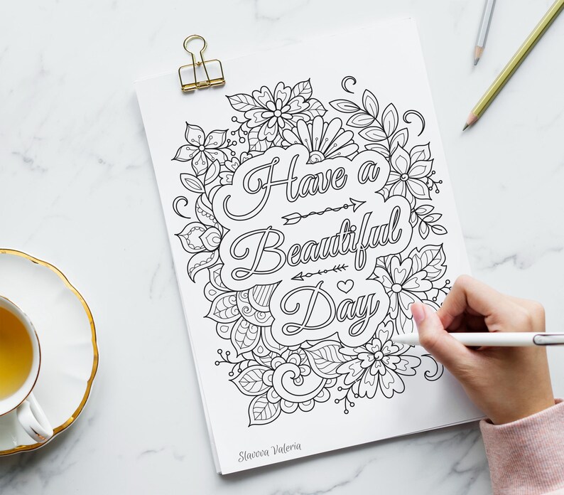 May include: A black and white floral coloring page with the words "Have a Beautiful Day" is held by a gold binder clip. A hand is coloring the page with a white pen. A cup of tea sits on a white saucer with a gold rim.