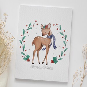 May include: A watercolor illustration of a fawn wearing a blue scarf and a holly wreath on its head. The fawn is standing in a wreath of green leaves and red berries. The text "Slavova Valeria" is written below the fawn.