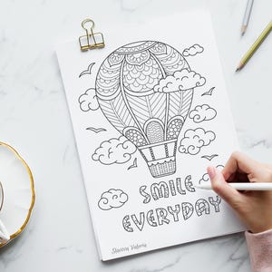 May include: Black and white line art of a hot air balloon, clouds, and birds on a white paper. The words "SMILE EVERYDAY" are written below the balloon. A hand is holding a pen and coloring the image. A gold paper clip holds the paper.
