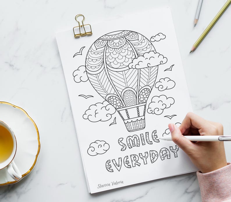 May include: Black and white line art of a hot air balloon, clouds, and birds on a white paper. The words "SMILE EVERYDAY" are written below the balloon. A hand is holding a pen and coloring the image. A gold paper clip holds the paper.