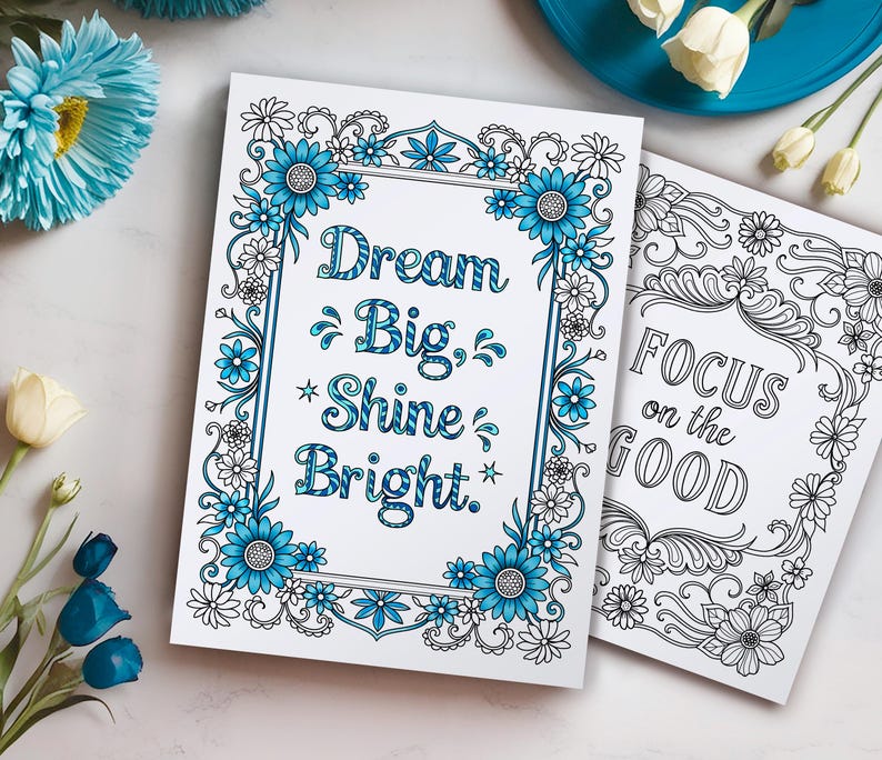 May include: Two printable coloring pages with inspirational quotes. The first page features a floral border with blue flowers and the quote "Dream Big, Shine Bright." The second page features a black and white floral border with the quote "Focus on the Good."