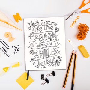 May include: A black and white printable coloring page with the text "Be the reason someone smiles today" surrounded by flowers. The page is on a white surface with various office supplies, including pencils, a rubber eraser, paper clips, and a pair of scissors.