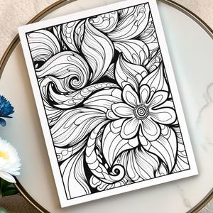 May include: A black and white floral line art print featuring a large flower and swirling leaf-like patterns. The artwork is on a white paper, resting on a marble-like surface. The print is suitable for coloring or as a decorative piece.