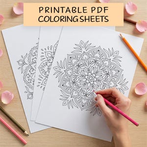 May include: Three printable colouring sheets featuring intricate mandala designs. The sheets are white with black outlines and are stacked on a wooden surface. A hand is holding a pink pencil and colouring one of the mandalas.