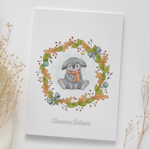 May include: A watercolor illustration of a gray bunny wearing an orange scarf sitting inside a wreath of green leaves, brown acorns, and red berries. The wreath is decorated with blue bows and gold ornaments. The text "Slavova Valeria" is written below the wreath.