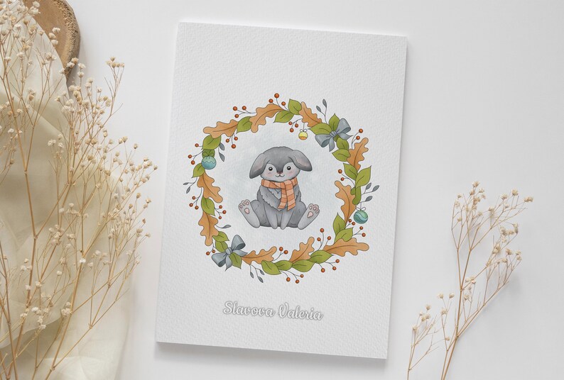 May include: A watercolor illustration of a gray bunny wearing an orange scarf sitting inside a wreath of green leaves, brown acorns, and red berries. The wreath is decorated with blue bows and gold ornaments. The text "Slavova Valeria" is written below the wreath.