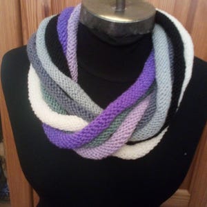 Pu&ograve; includere: Una sciarpa infinita di maglia multicolore con sfumature di viola, bianco, grigio e nero. La sciarpa &egrave; esposta su un manichino nero.