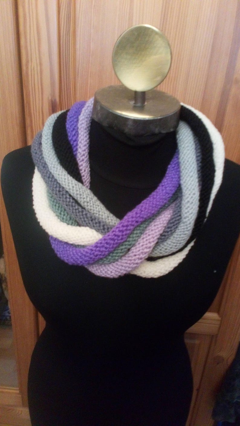 Pu&ograve; includere: Una sciarpa infinita di maglia multicolore con sfumature di viola, bianco, grigio e nero. La sciarpa &egrave; esposta su un manichino nero.